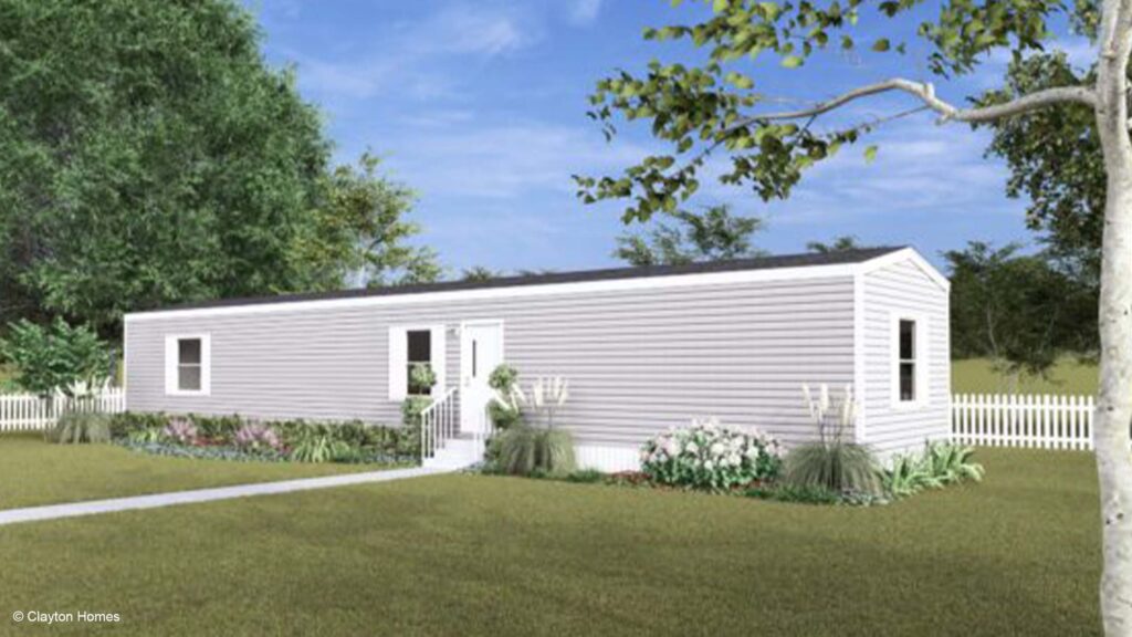 A white manufactured home with a small garden and white picket fence sits under a blue sky, with trees in the background.