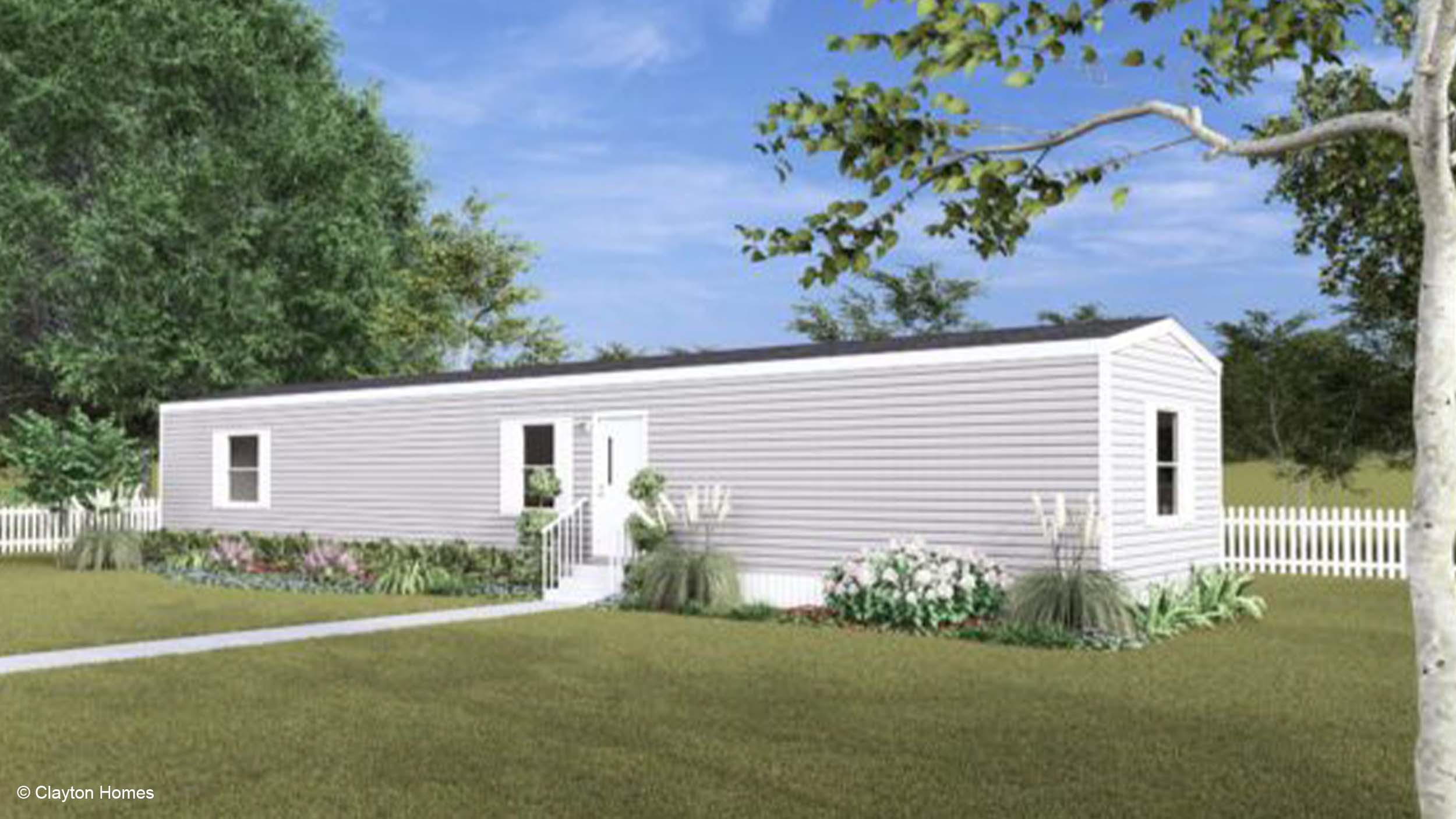 A white manufactured home with a small garden and white picket fence sits under a blue sky, with trees in the background.